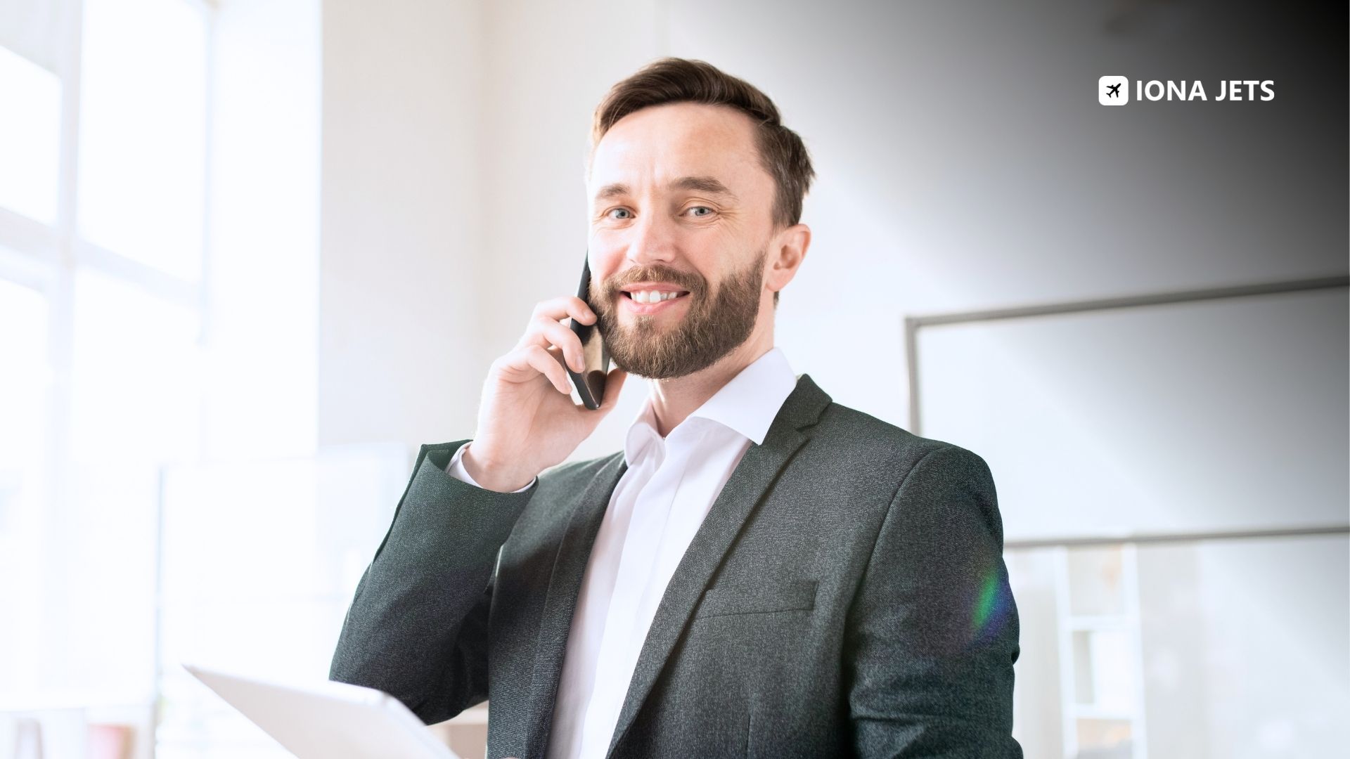 Charter broker in a business suit smiling while taking a phone call and reviewing flight documents at the office, coordinating a private jet charter request with operator partners.